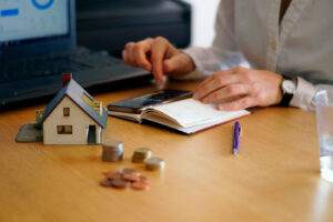 Foto em close de uma pessoa utilizando a calculadora e com uma maquete de casa e algumas moedas sobre a mesa